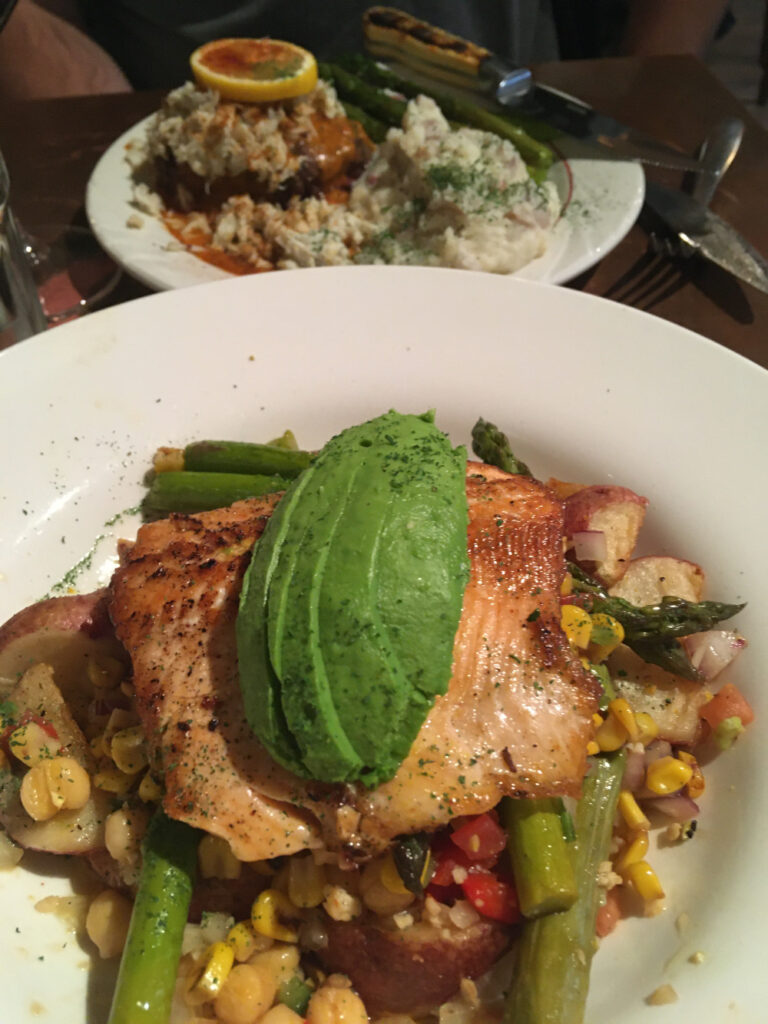 A plate filled with a base of vegetables topped with trout topped with an avocado