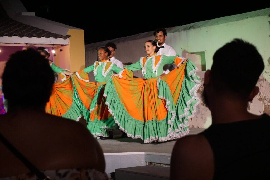 Female dancers dressed in colorful dresses of green and orange, holding out their skirts. Men dressed in white shirts and black bow ties in the background.