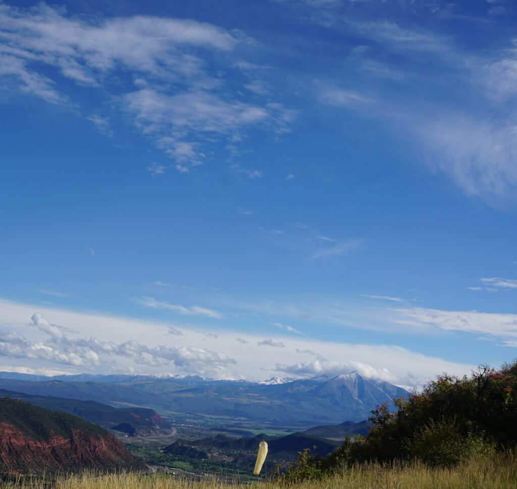 This photo shows the town of Glenwood Springs, Colorado, from the top of a mountain hiking trail. The sky is blue with a few clouds and there are mountains in the distance. The town is nestled in a valley.