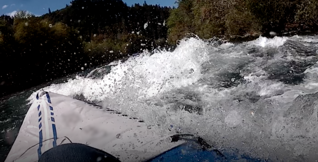 This photo shows the front of end of a kayak in a river rapid. The kayak tilts to one side. There are trees in the background.