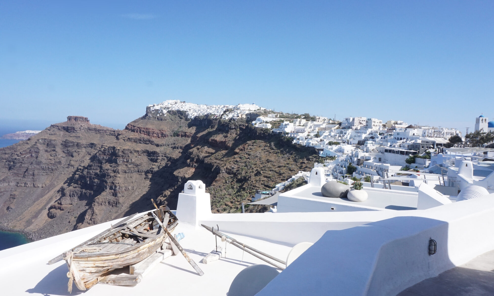 In the foreground old wooden boat in on top a white-washed building. In the background, white-washed building stretching to the edge of a cliff, the town of Oia on the island of Santorini, Greece.