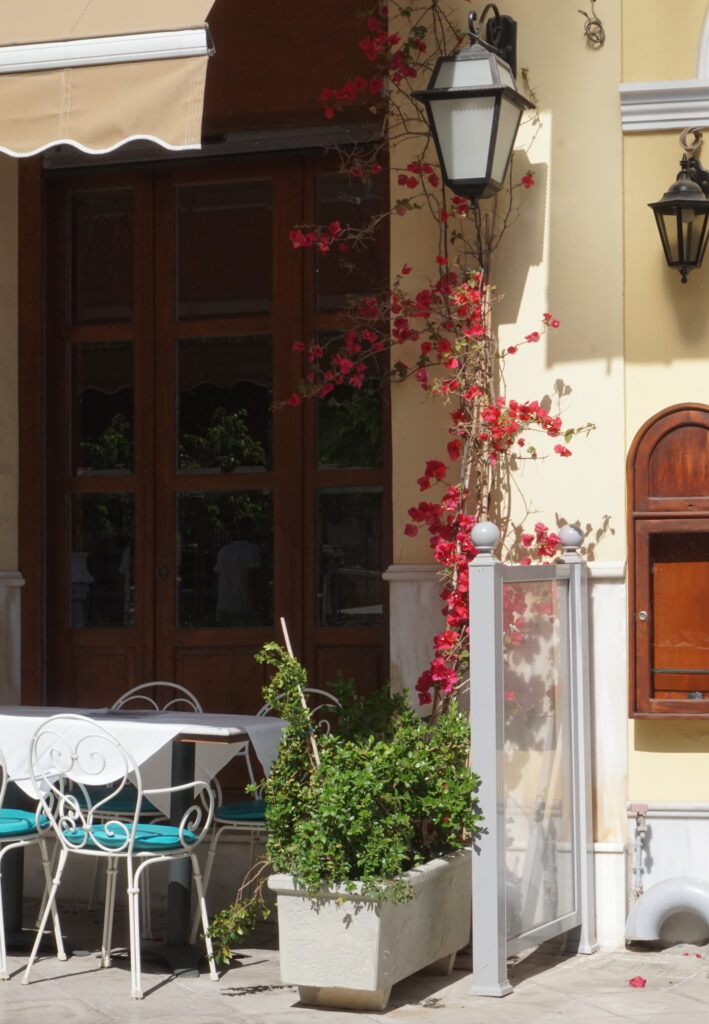 Exterior of a restaurant with white-cloth-covered table and white metal chairs with aqua seats. Pink Bougainvillea climbs the wall.